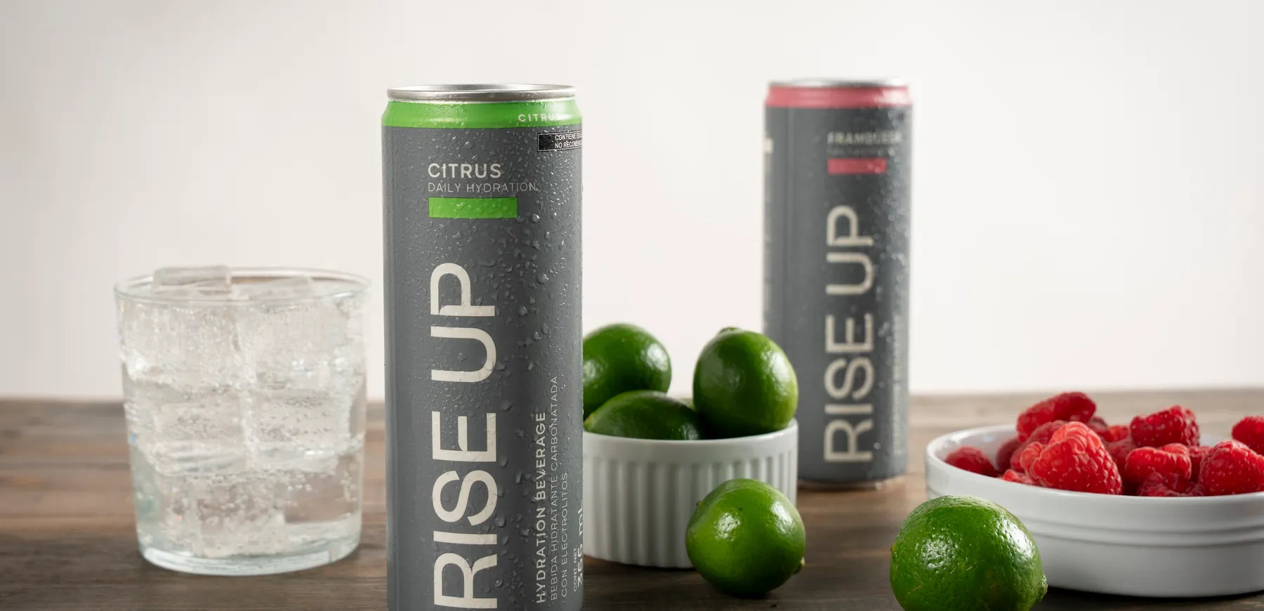 Latas Rise Up Citrus y Frambuesa sobre una mesa de madera, con limones y un vaso con hielo